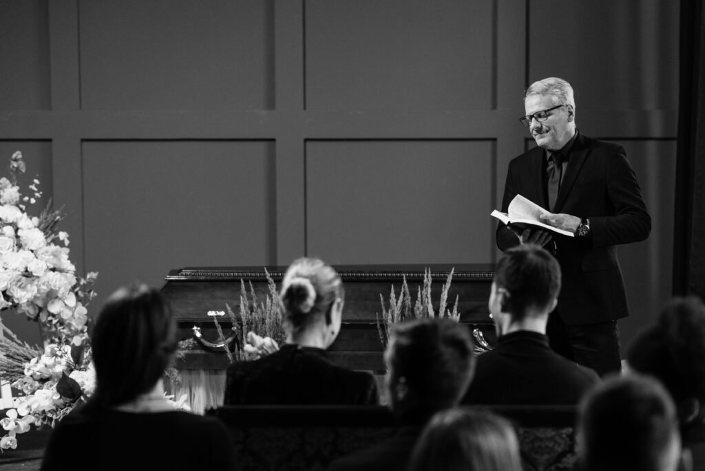 A solemn funeral ceremony indoors with attendees and a pastor speaking.